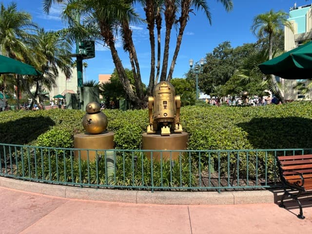 Two bronze droid statues on pedestals surrounded by hedges at Walt Disney World, with tall palm trees and park buildings in the background