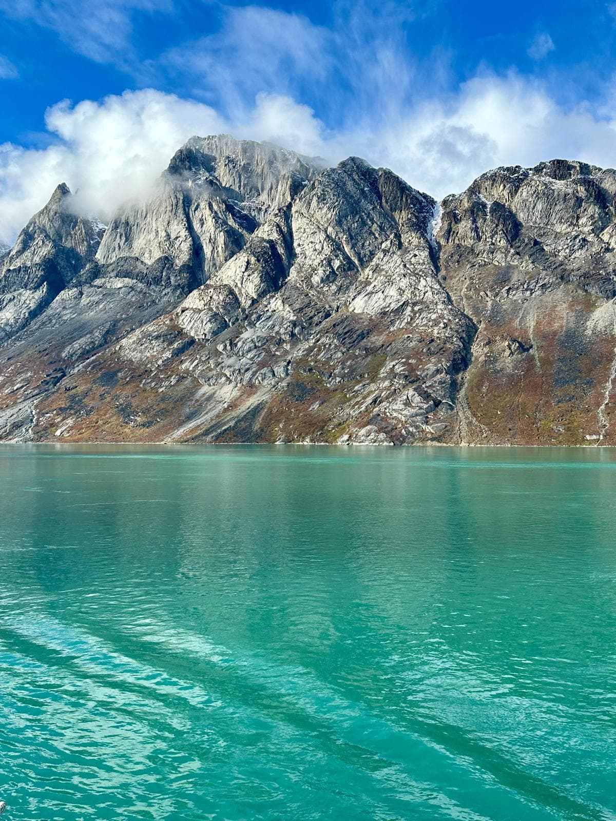 Dramatic rocky peaks rise above turquoise glacial water under a blue sky with white clouds in Greenland