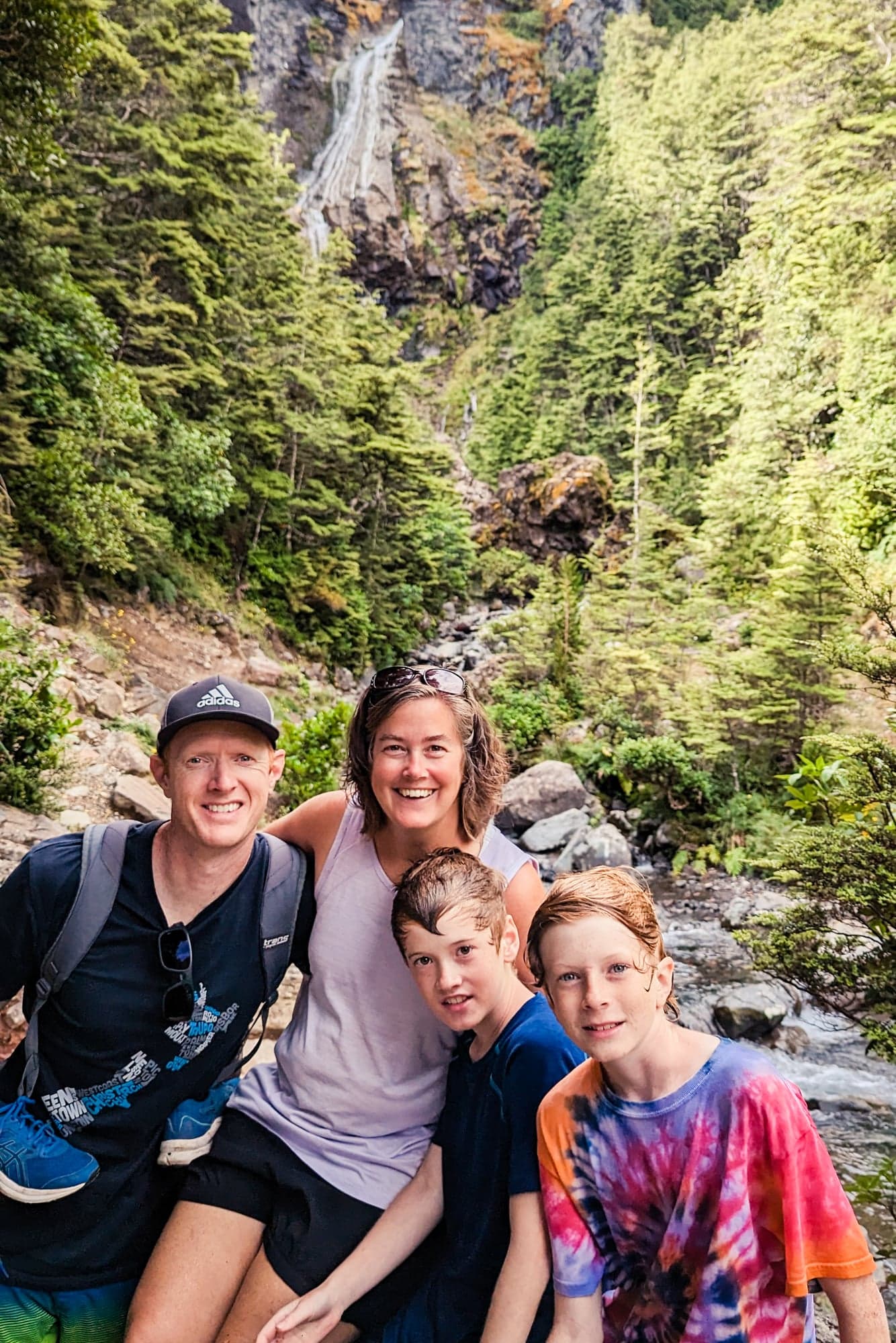 Family of four smiling at a stream in a lush Costa Rica canyon with forested cliffs and a waterfall visible above