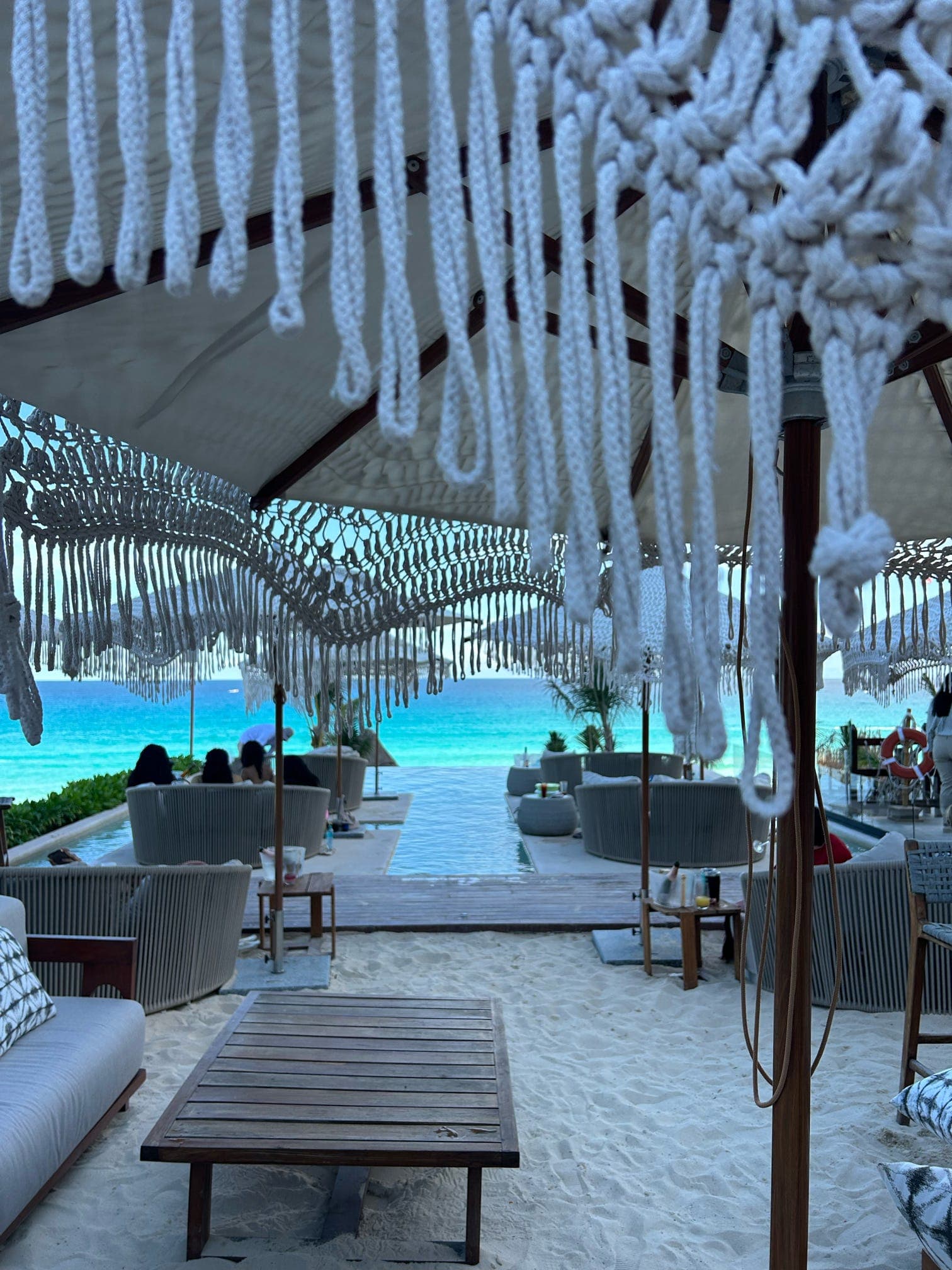 White macramé curtains frame a beachfront lounge area with turquoise ocean and palm trees visible beyond