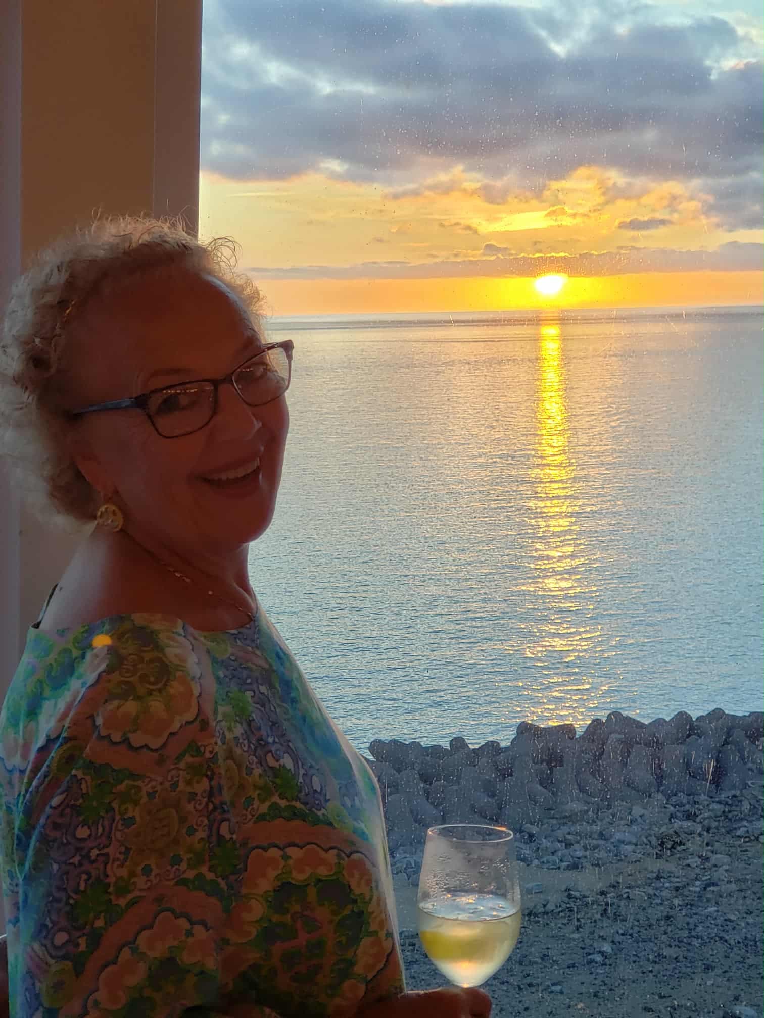 Patti Carter smiling at sunset over calm ocean waters, holding a glass of white wine on a rocky shore