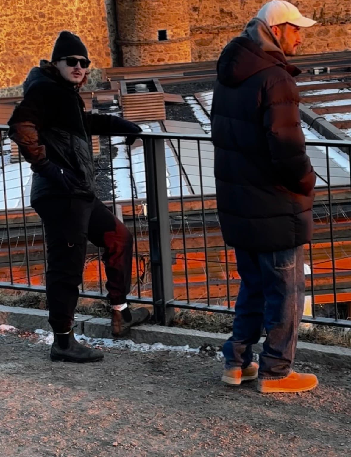 Two men standing on a wooden platform against a stone fortress wall in winter