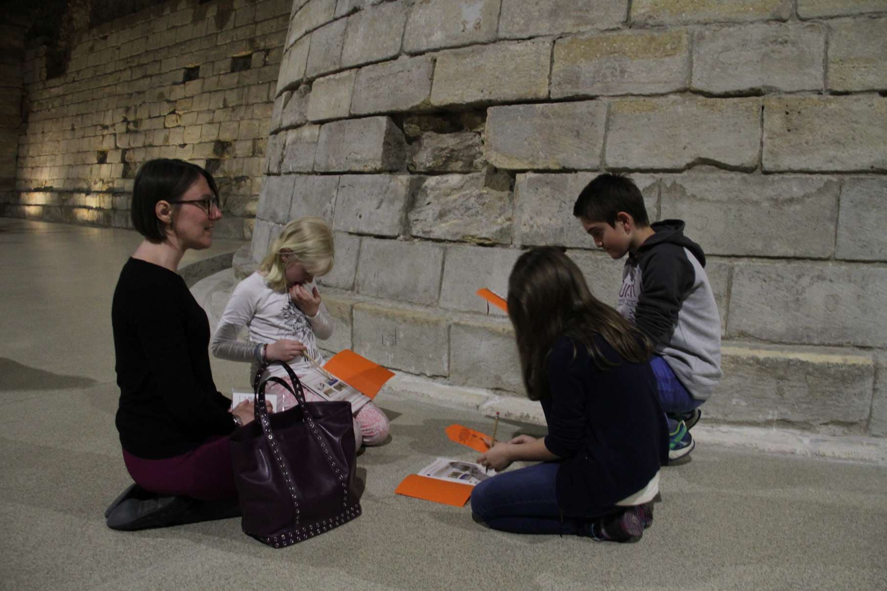 An adult and three children sit on a stone floor against ancient stone walls, examining orange educational materials together