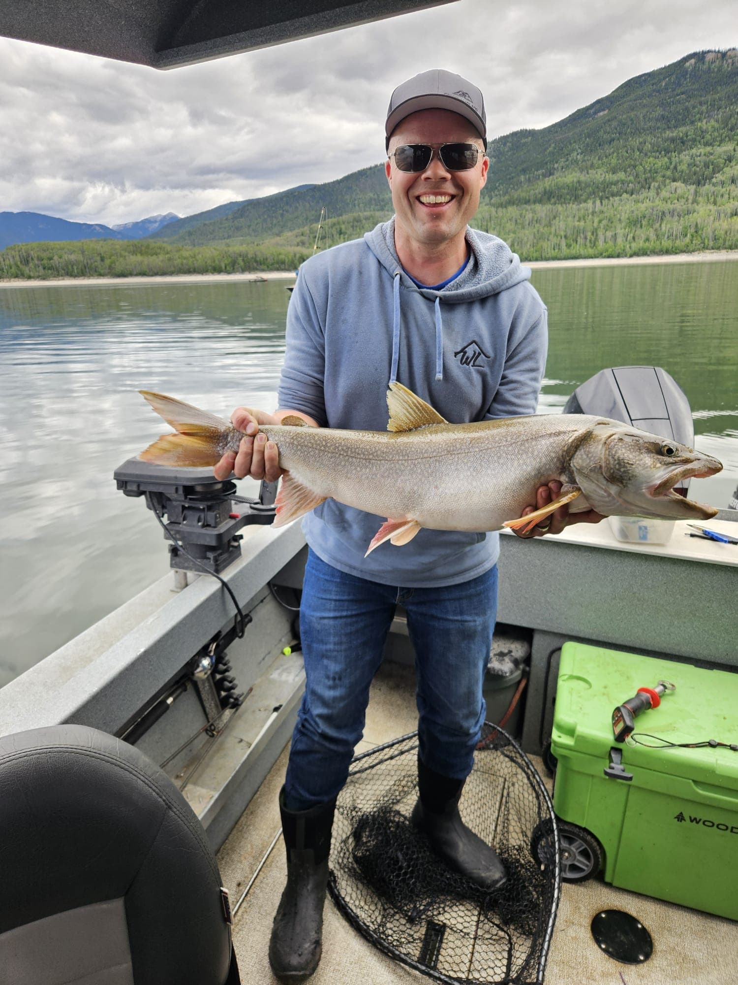 Lois Hirtz holds a large salmon on a fishing boat in Norway, with forested mountains and a calm lake in the background