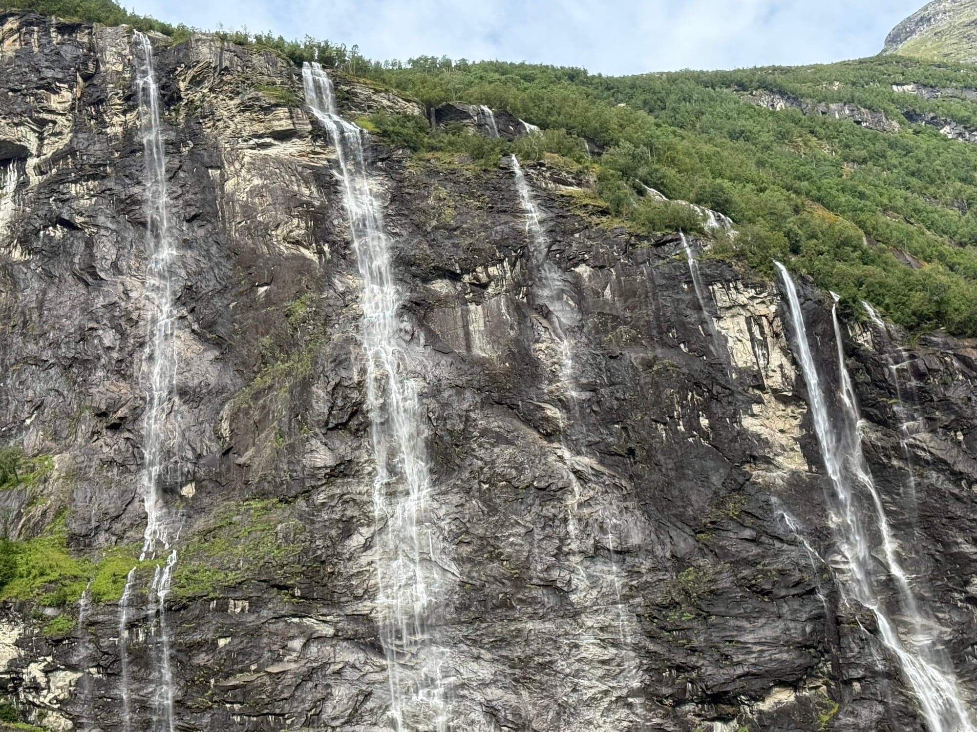 Multiple thin waterfalls cascade down a steep rocky cliff face surrounded by green forest in Norway
