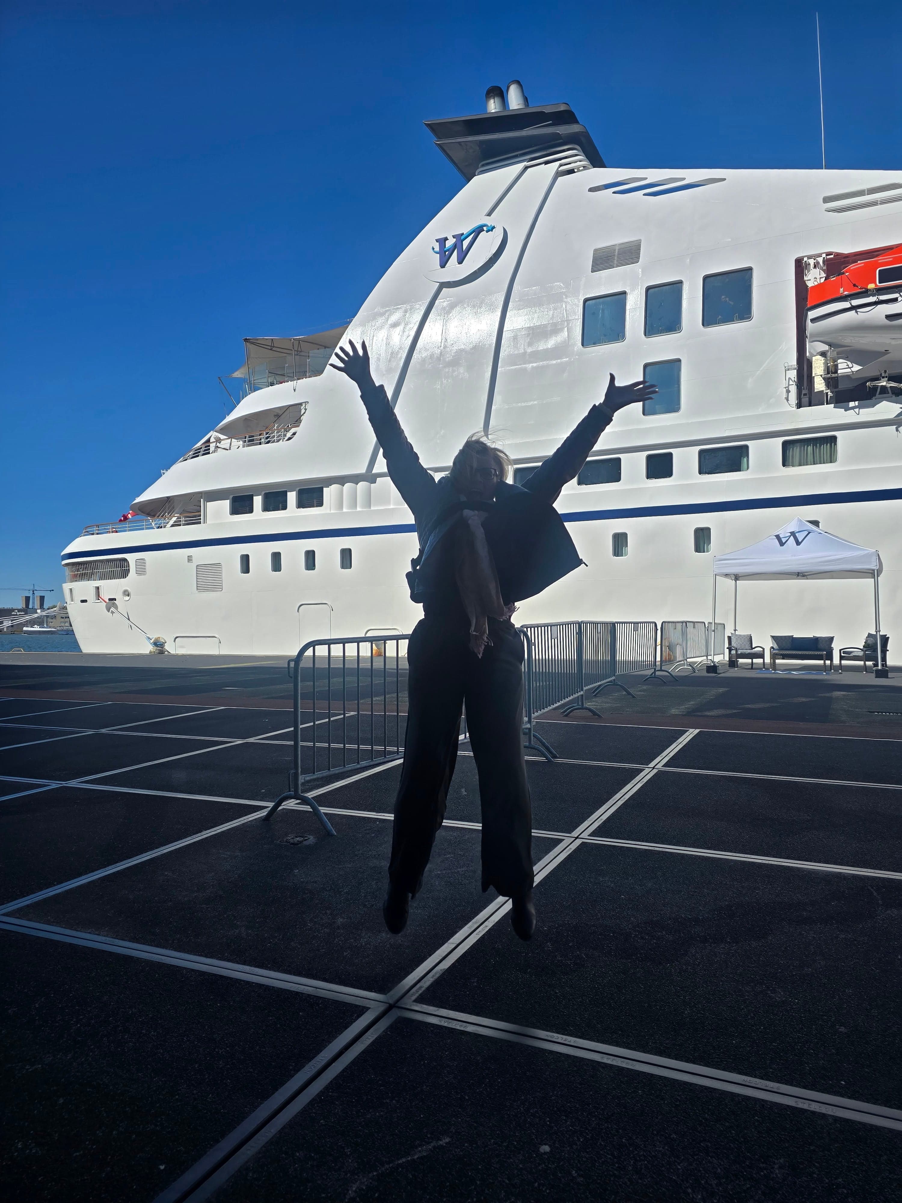 Woman in dark clothing jumping with arms raised in front of a large white cruise ship docked at port