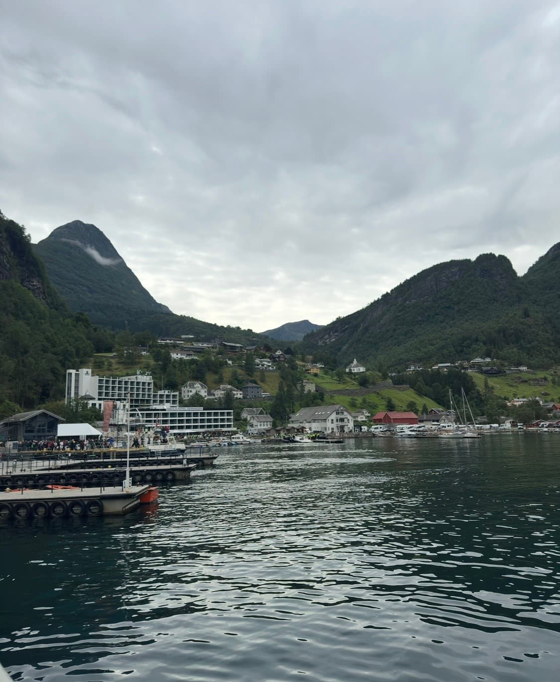 Cruise ship docked at a fjord port with steep mountains and modern hotel buildings in the background under cloudy skies