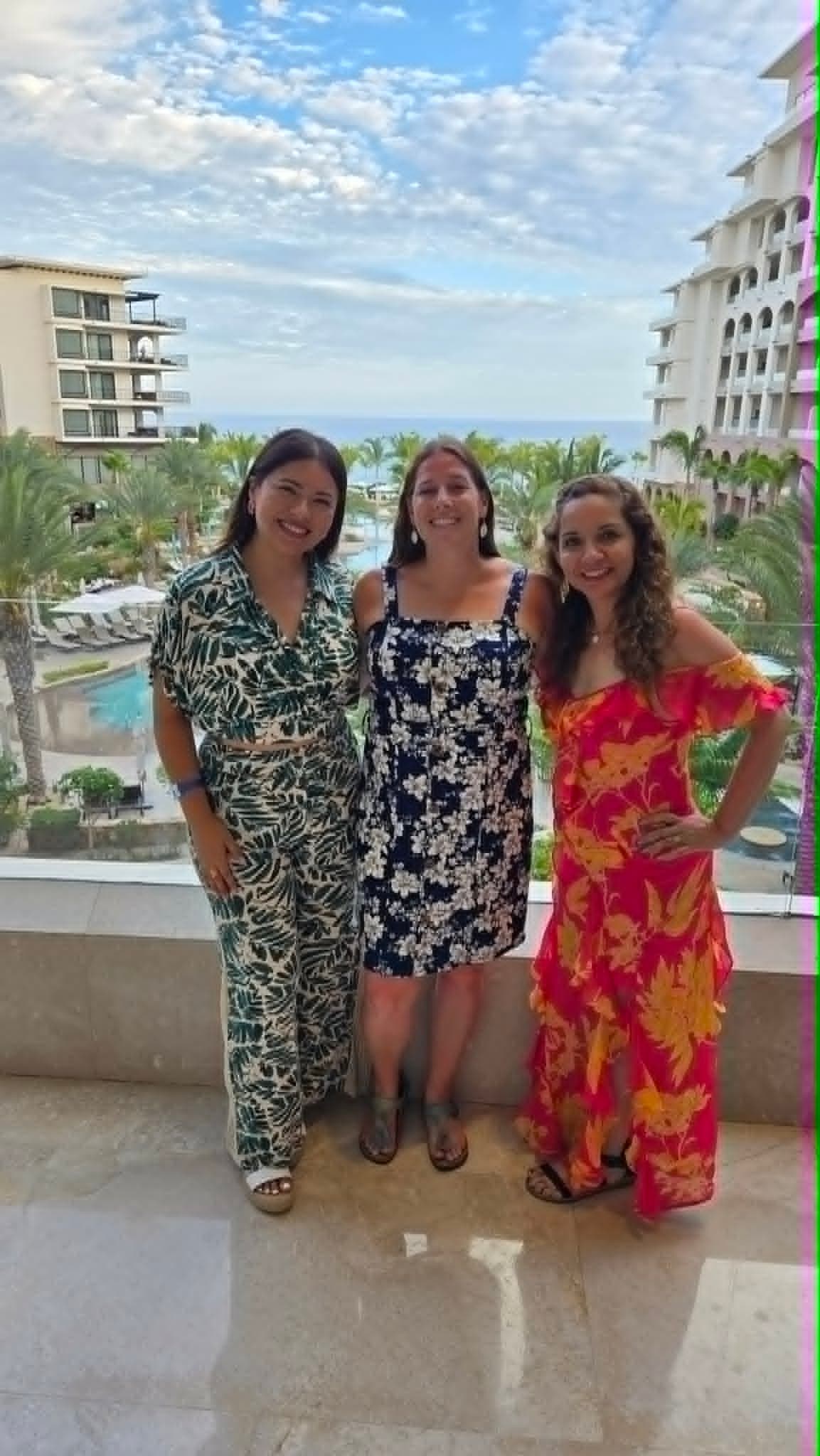 Three women in colorful patterned dresses pose on a resort balcony overlooking palm trees and the ocean in Los Cabos