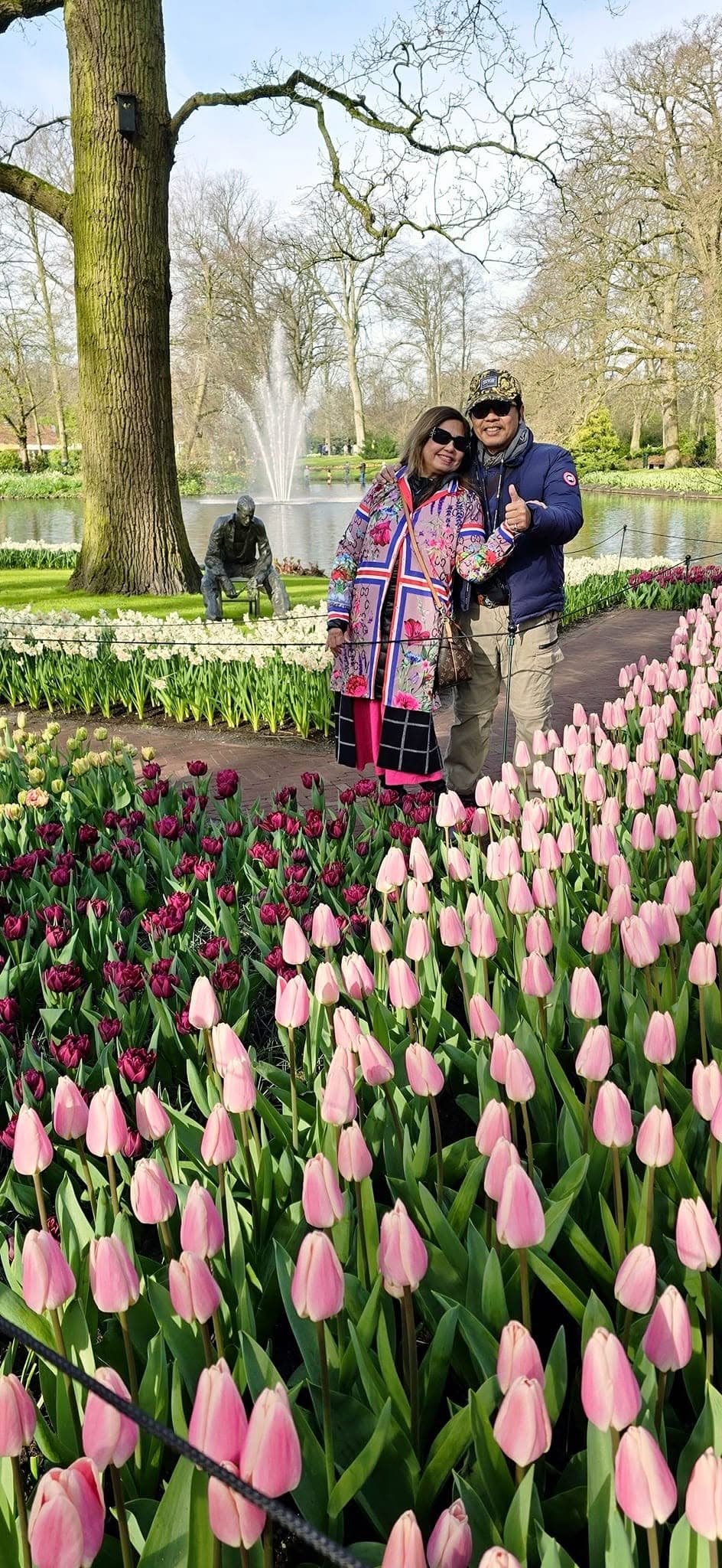 Cleofe Cornelio and companion posing among pink and purple tulips in a Netherlands garden with fountain and bare trees