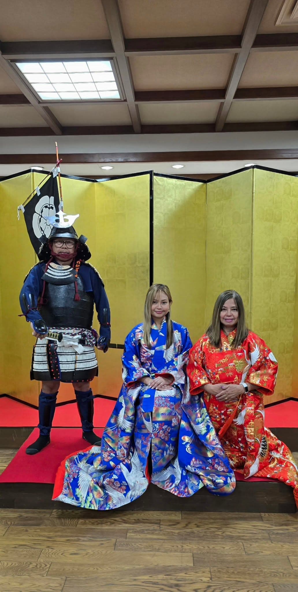 Cleofe Cornelio in samurai armor costume posing with two women in colorful kimono against yellow backdrop