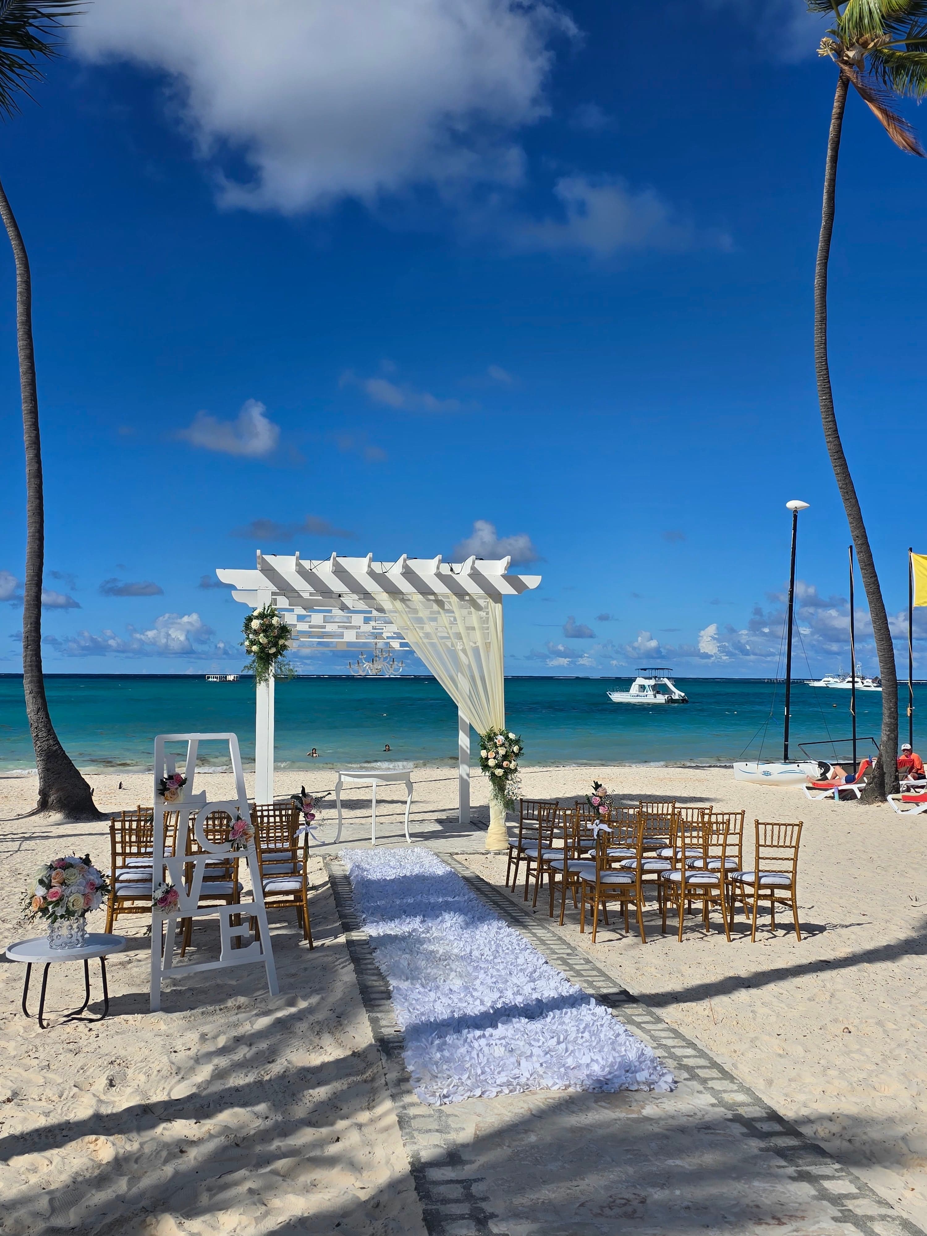 White wedding ceremony setup on a sandy beach in Punta Cana with a pergola, white aisle runner, and turquoise ocean under blue sky