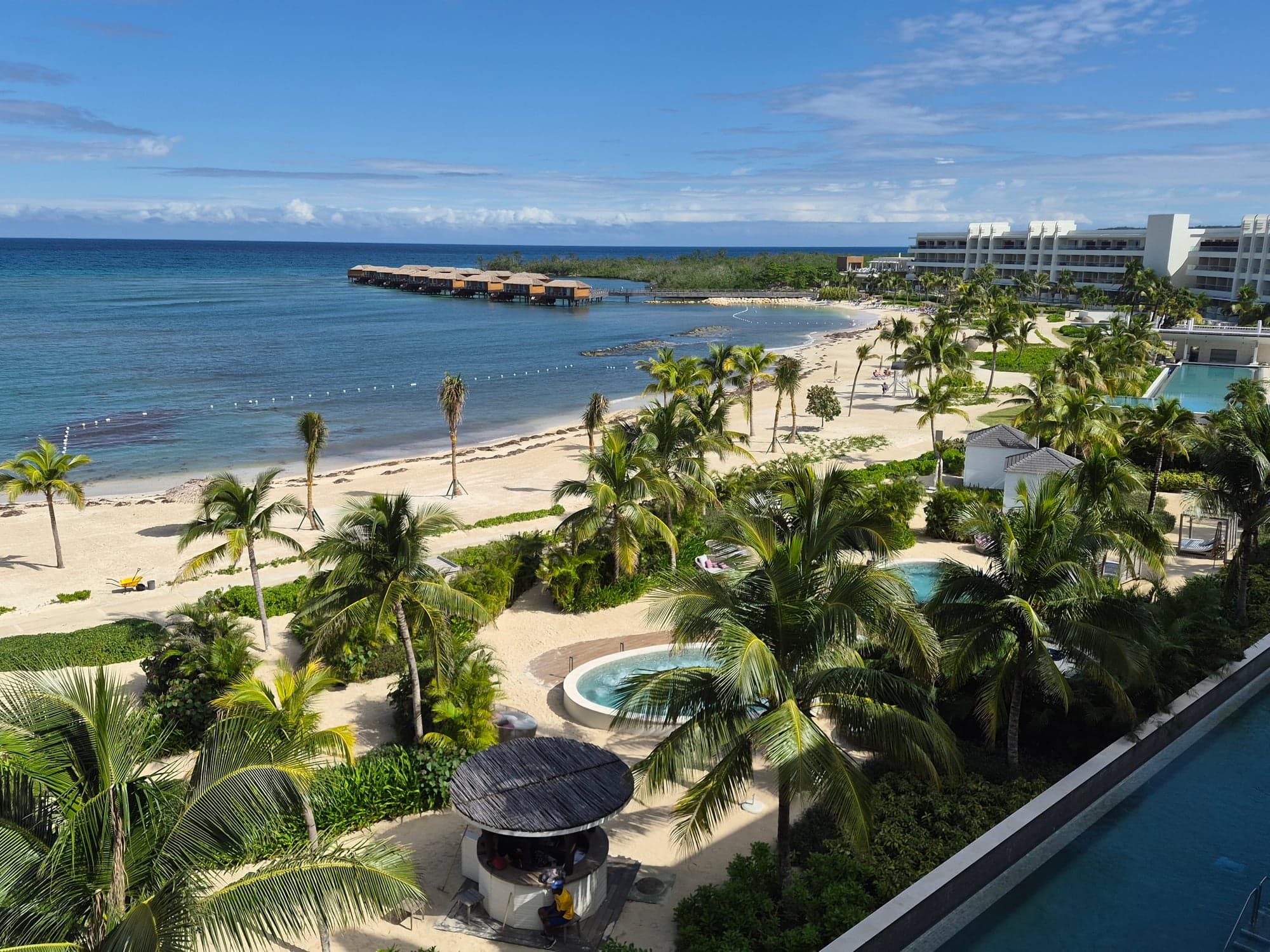 Aerial view of Princess Senses Jamaica resort with white buildings, palm trees, beach, and turquoise ocean with overwater structures