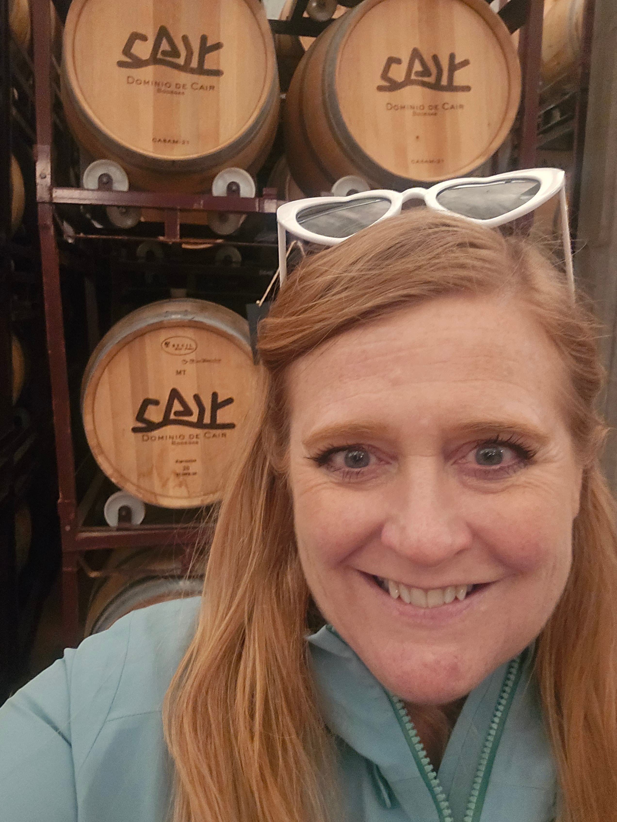 Crystal Sturgis smiles in front of wooden wine barrels labeled 'Cair' at a Spanish winery