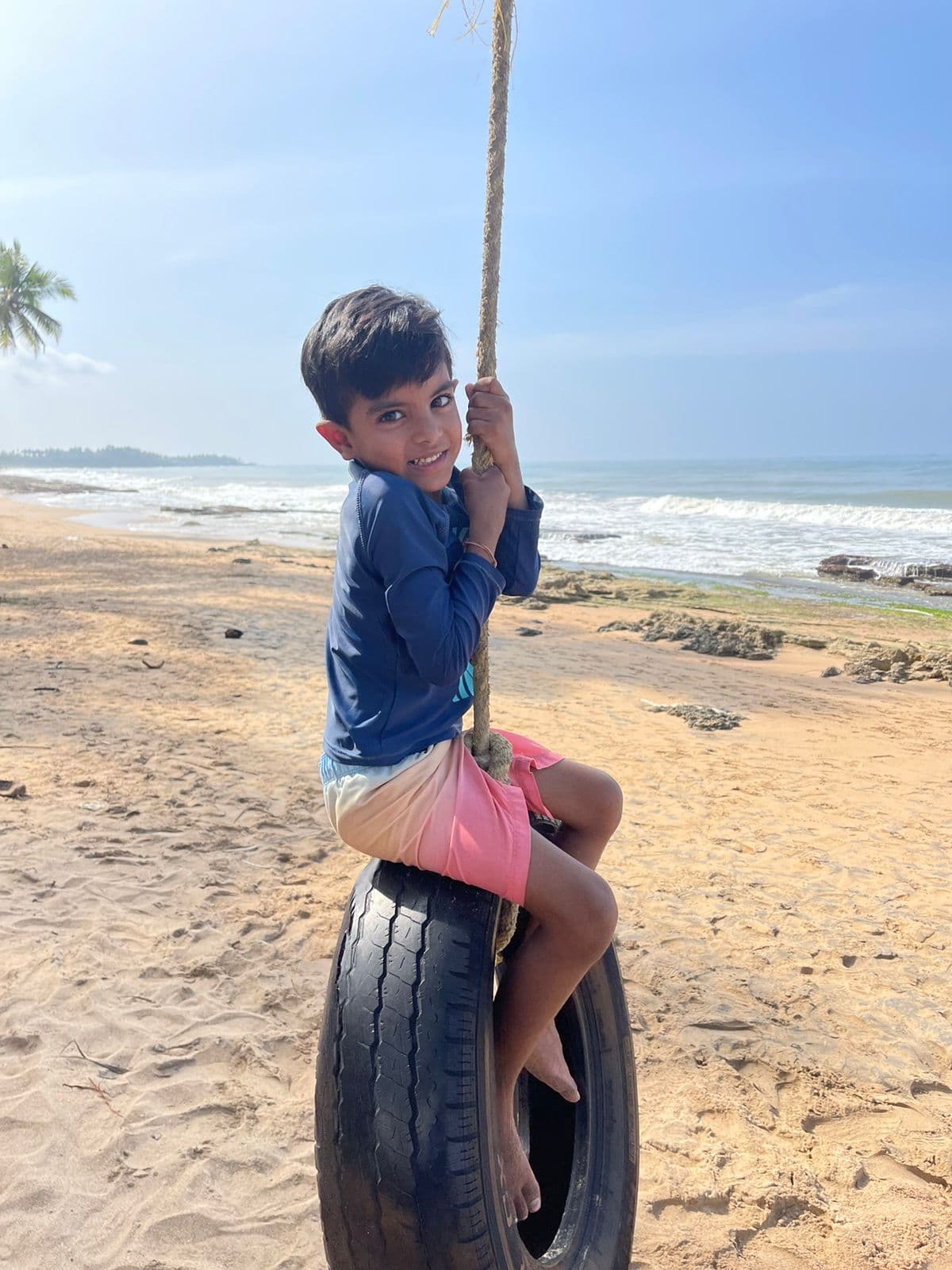 Young boy smiling while holding a rope swing attached to a palm tree on a Sri Lankan beach with waves in the background