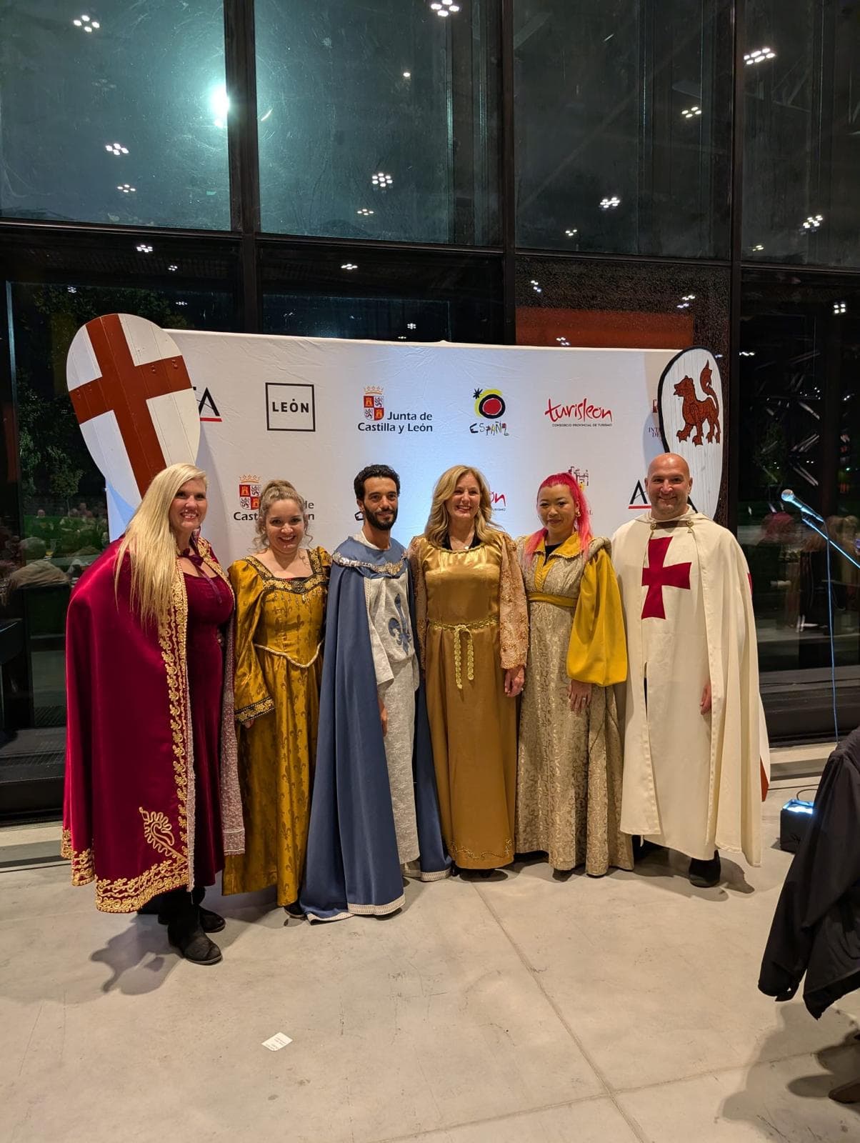 Six people in medieval costumes stand before a backdrop with Castilla y León regional logos and symbols at a nighttime event