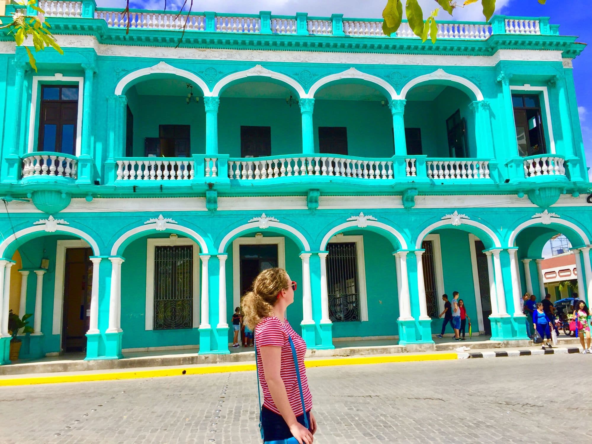 Autumn Fiske stands before a vibrant turquoise colonial building with white arched columns and balustrades in Cuba