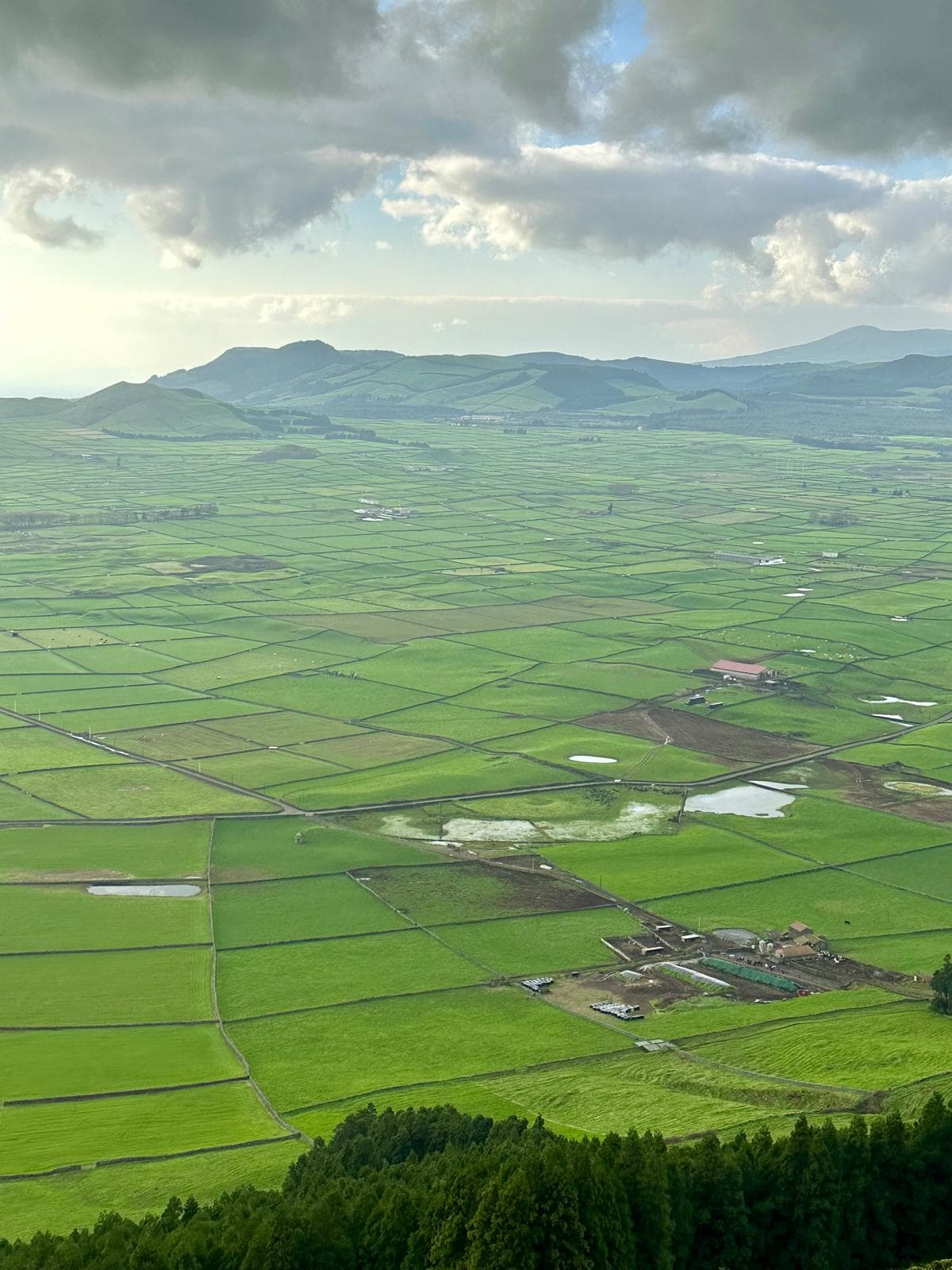 Aerial view of patchwork green agricultural fields with distant mountains under cloudy sky in the Azores