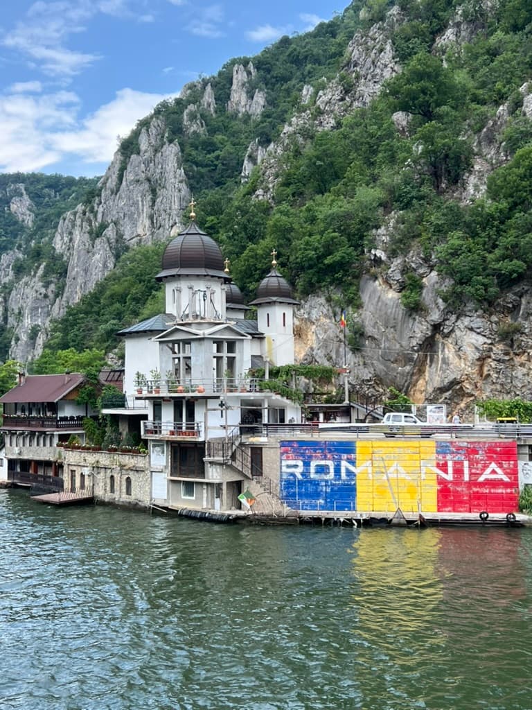 White Orthodox church with black domes nestled at the base of dramatic limestone cliffs along the Danube River in Romania