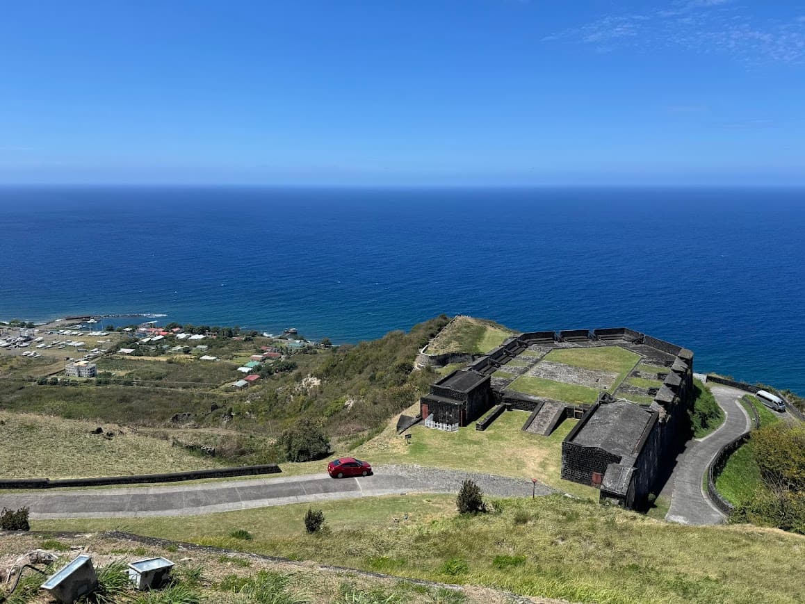 Coastal fortress with black stone buildings on grassy headland overlooking blue ocean and small settlement below