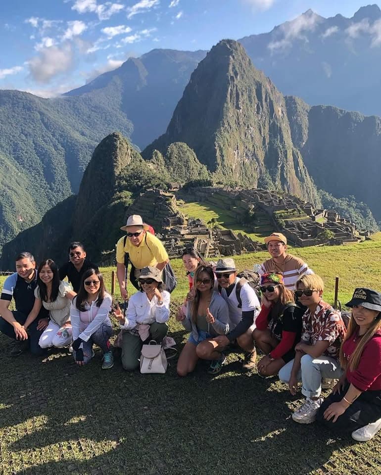 Large group of tourists posing together on grass with Machu Picchu's iconic peak and terraces visible in the background