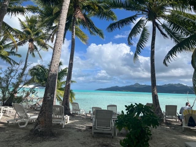 Beach lounge chairs beneath palm trees facing turquoise lagoon and distant volcanic islands in Tahiti