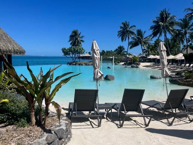 Turquoise lagoon with white umbrellas and lounge chairs on a sandy beach lined with palm trees in Tahiti