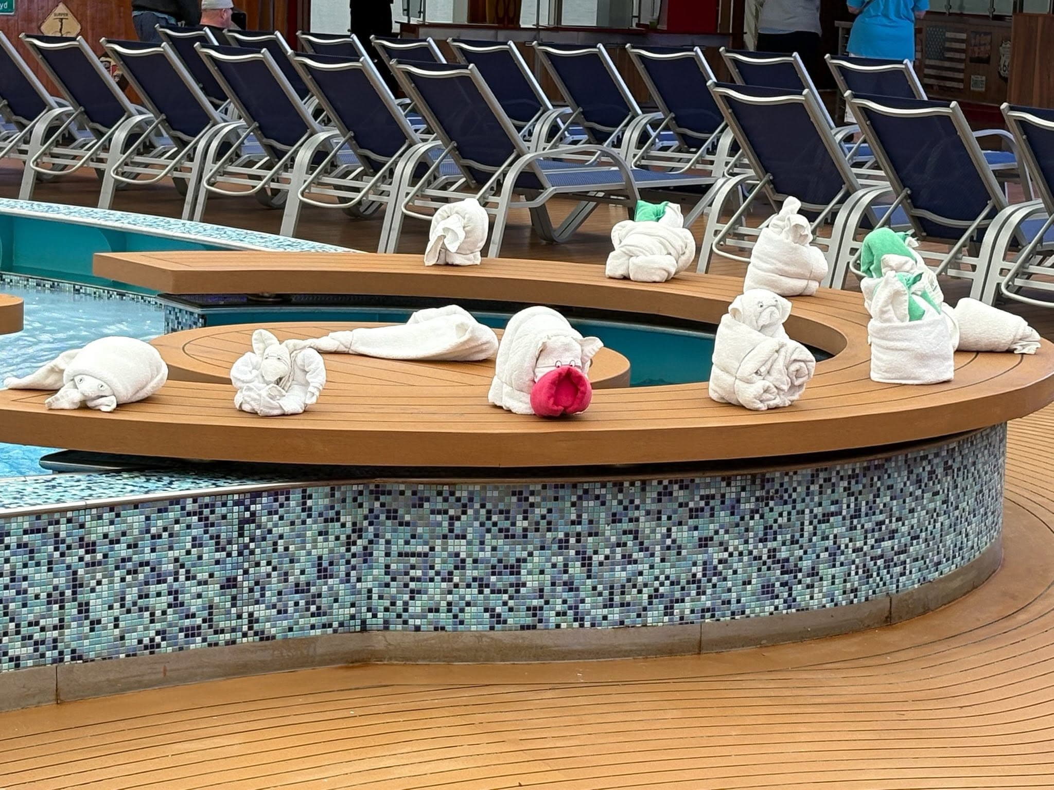 Poolside lounge chairs with white rolled towels arranged on wooden deck surrounding a curved hot tub at a cruise ship