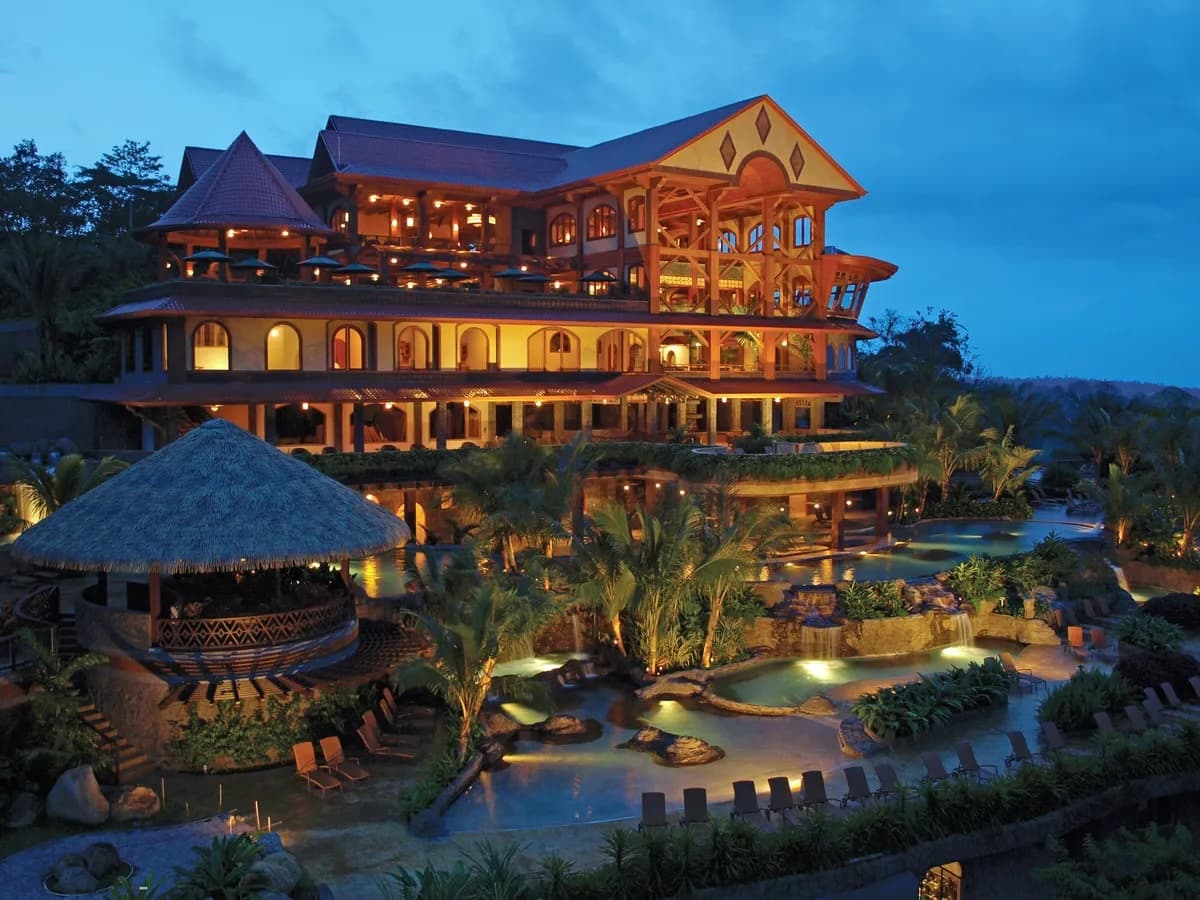 Illuminated multi-story lodge with distinctive architecture overlooks a glowing pool and tropical gardens at dusk in Arenal, Costa Rica