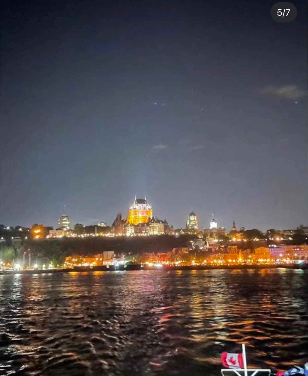 Nighttime view of Quebec City's skyline across the St. Lawrence River, with illuminated cathedral and historic buildings, Canadian flag visible in